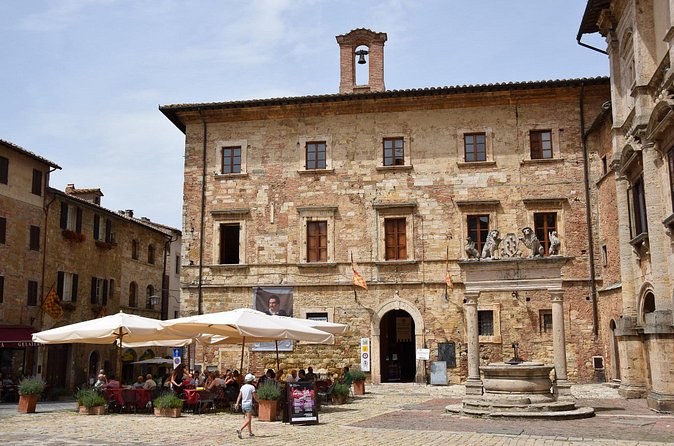 A palace front on Piazza Grande with café tables under umbrellas, and the columned well standing beside it